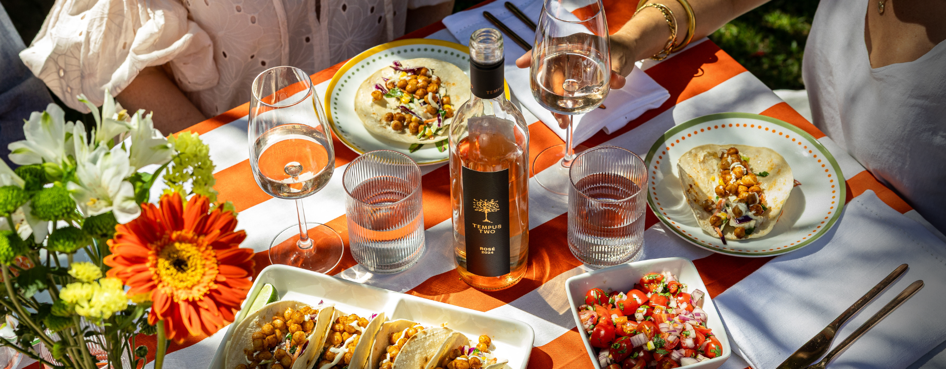 Summer table with chickpea tacis, orange and white striped table cloth Tempus Two varietal rose bottle and glasses poured with wine in the summer sun.