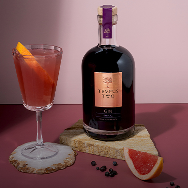 Bottle of Tempus Two Shiraz Gin on a slate of sandstone with a glass of Tempus Two shiraz gin, garnished with an orange
