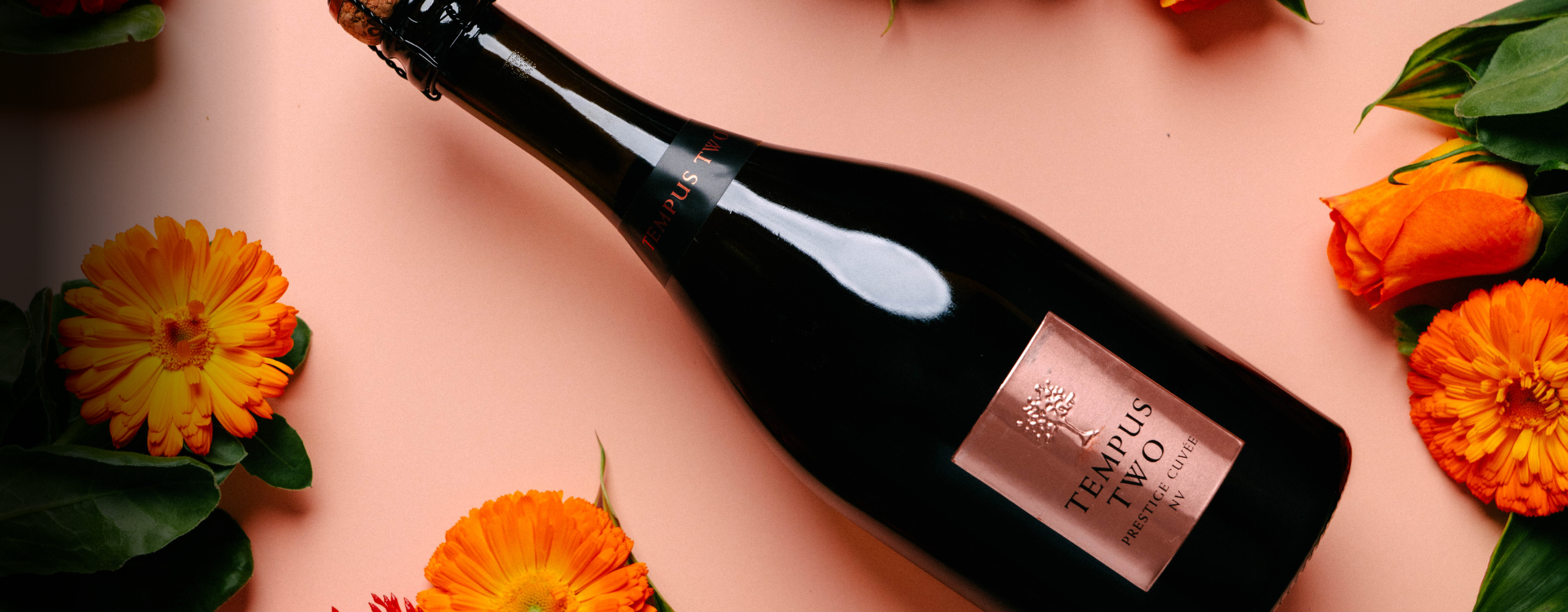 Tempus two Prestige cuvve bottle lying down on top of a pink backdrop and bright orange flowers